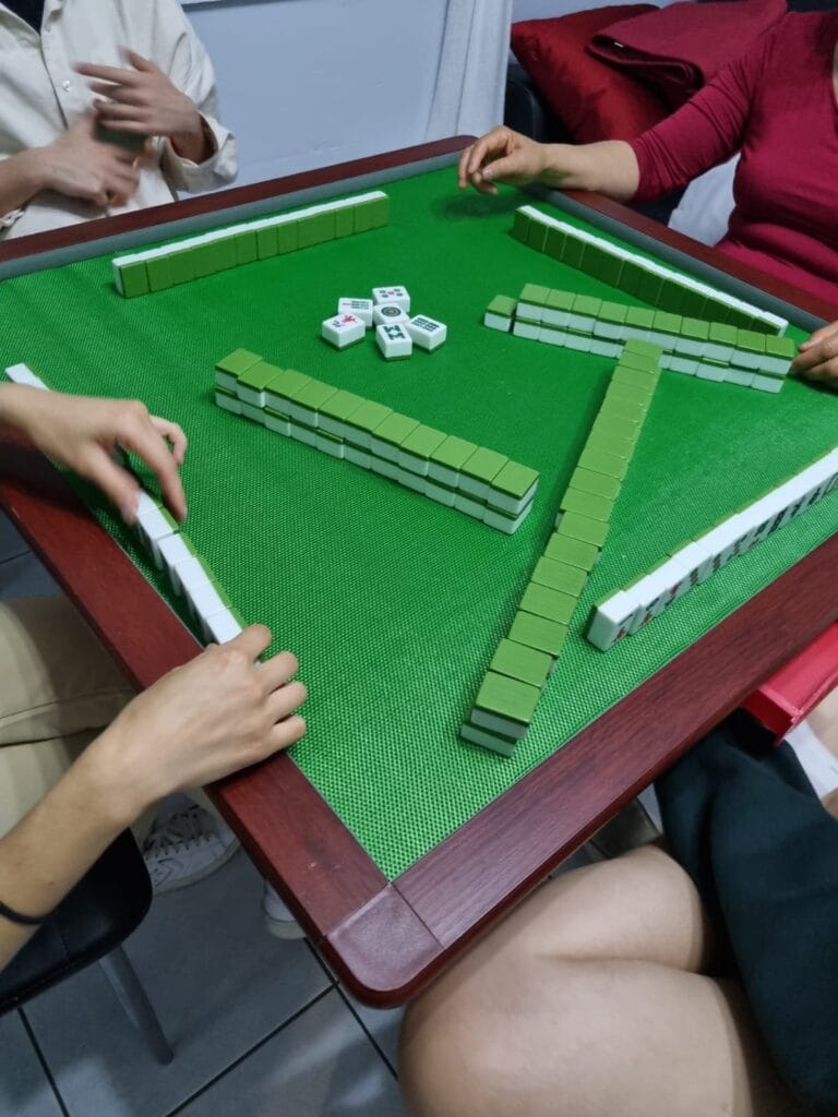Players reaching for tiles around a green mahjong table during a game