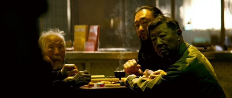 Film still of players gathered around a mahjong table in a dimly lit room